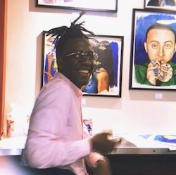 Dominique Barlow headshot, a black man wearing glasses and a pink shirt sitting at a desk with art behind him