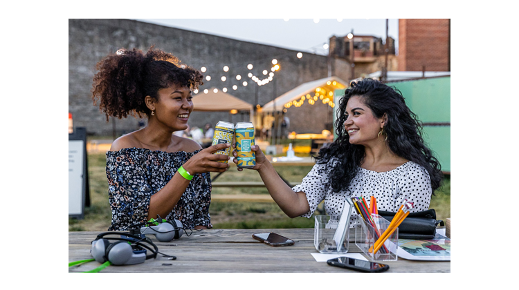 Two people holding cans of beer and smiling while sitting at a picnic table