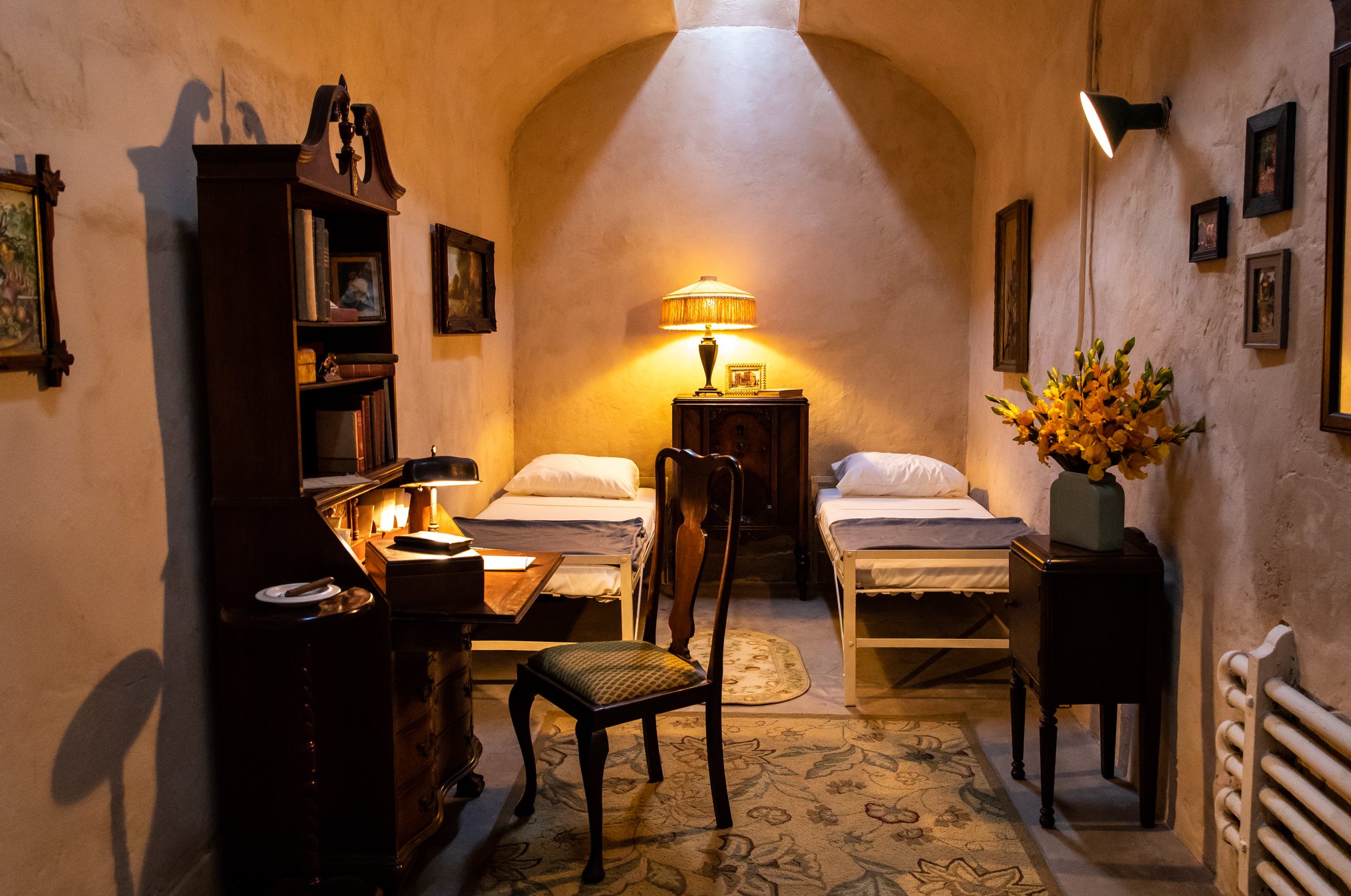 An image of AL Capone's cell at Eastern State Penitentiary. There are two single beds on a patterned rung. A wooden desk holds a lamp, books, and a cup. A small table nearby features a vase with yellow flowers. The walls are adorned with framed pictures and paintings.