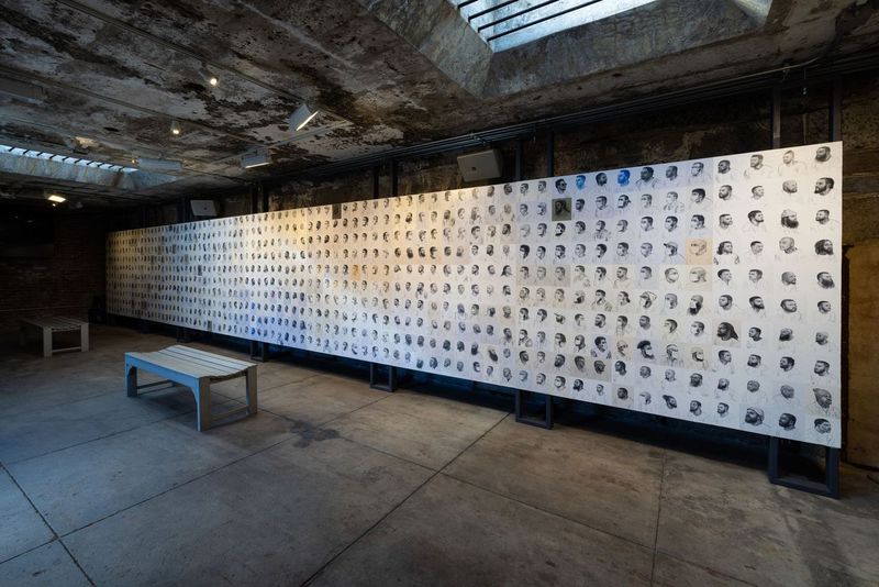A photo of artist Mark Loughney's exhibit Pyrrhic Defeat at Eastern State Penitentiary. The image shows a grid of hand-drawn shoulder-up portraits, each capturing individuals in varied poses—some facing forward, others in profile or three-quarter view.
