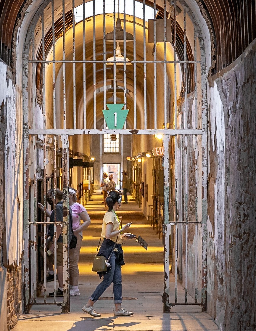 A photo of tourists at Eastern State Penitentiary, show a long, narrow cellblock with high arched ceilings, lined with rusted metal-barred cells on both sides. Several people walking through the corridor . A green sign with the number