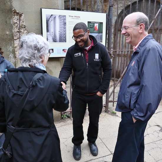 A photo of a tour guide talking to guests at Eastern State Penitentiary. The tour guide is shaking hands with one of the guests. Behind them, the display board features text and images related to
