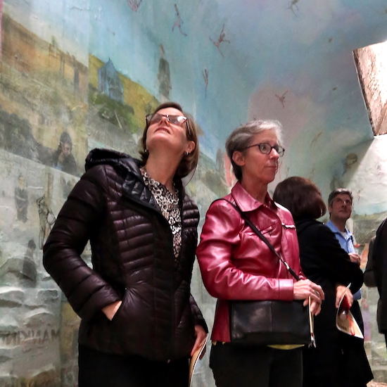 A photo of guests inside the cell with artist Jesse Krime's exhibit, Apokaluptein16389067:II. The cell walls and ceiling are covered in various photographs and images, creating a collage effect, which the visitors are observing.