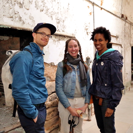 A photograph of guests at Eastern State Penitentiary. This image shows three individuals standing in a cellblock with peeling paint and exposed brickwork.