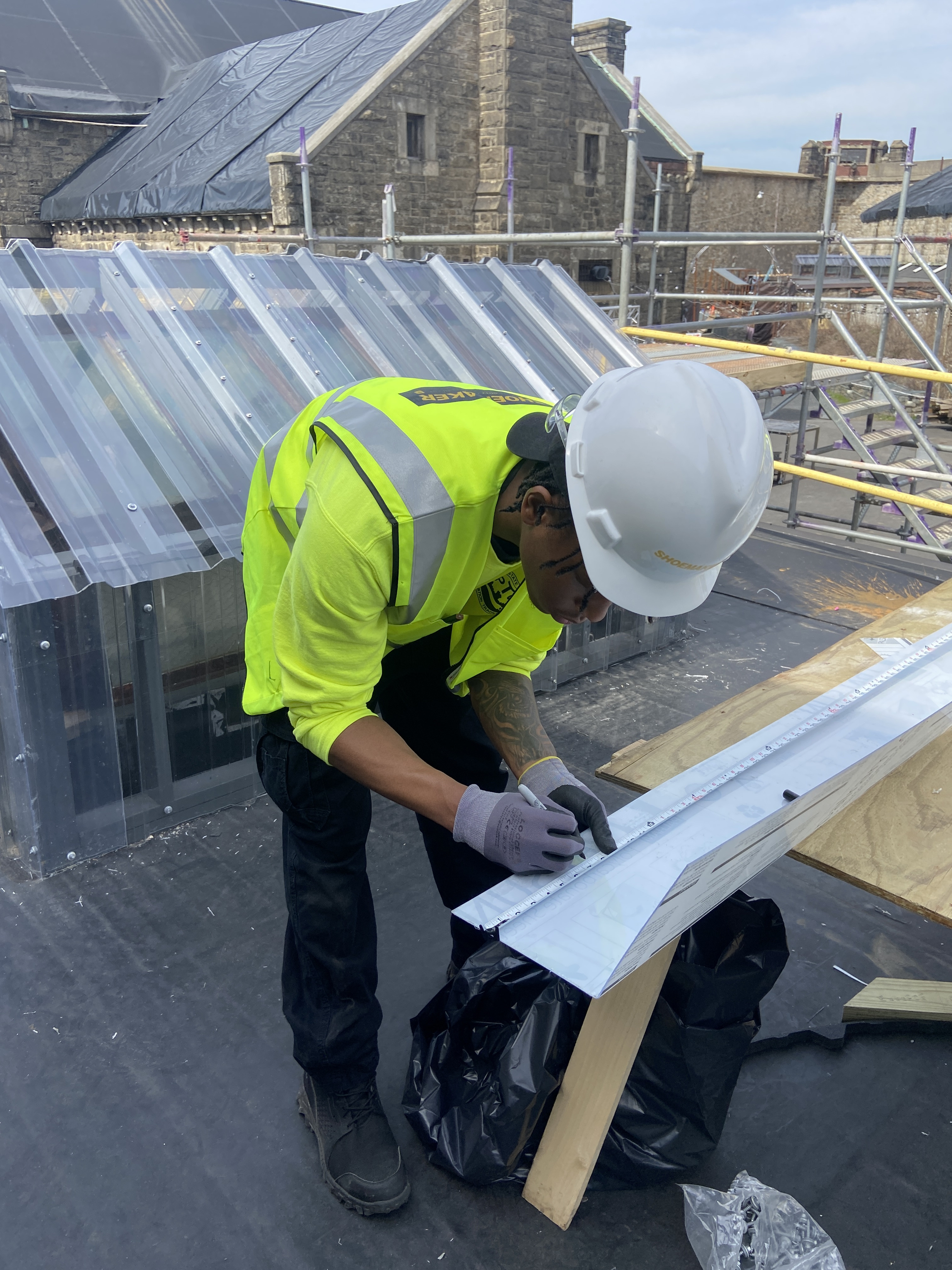 A photo of a LEAD fellow at Eastern State Penitentiary. They are wearing a hard hat, a safety vest, and marking off a board for cutting. A large stone building and the exterior wall are visible in the background.