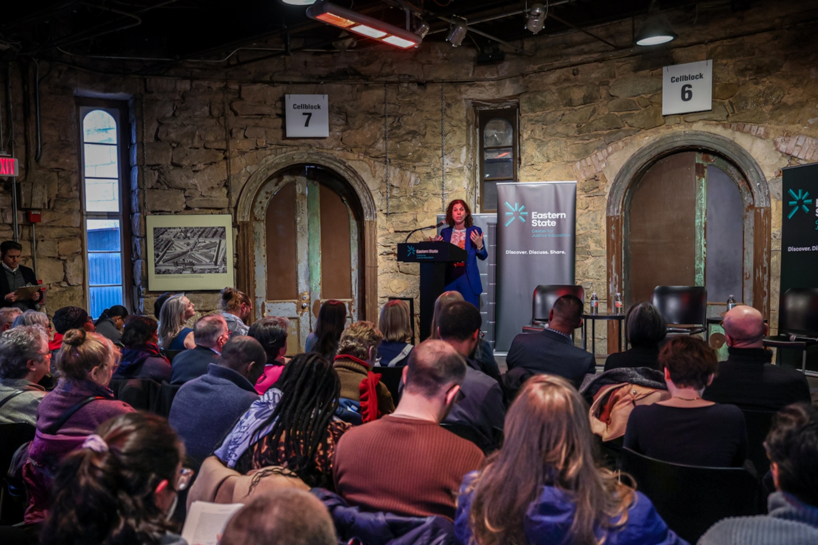 A person in a suit with dark curly hair stands at a podium on a stage in the center of Eastern State Penitentiary Historic Site while an audience looks on.