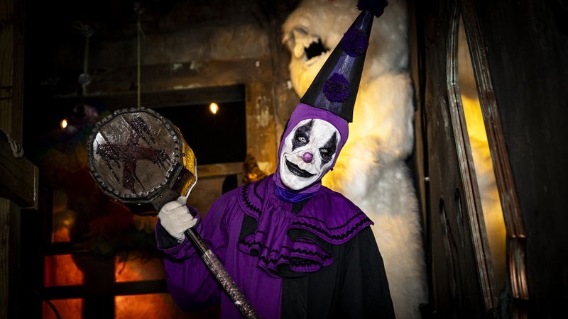 A person in a clown costume wears a purple and black jester outfit, a tall conical hat, and white face paint with black around the eyes and mouth. They hold a large mallet with a blood-stained head. A large, white, furry monster figure is visible in the dark background.