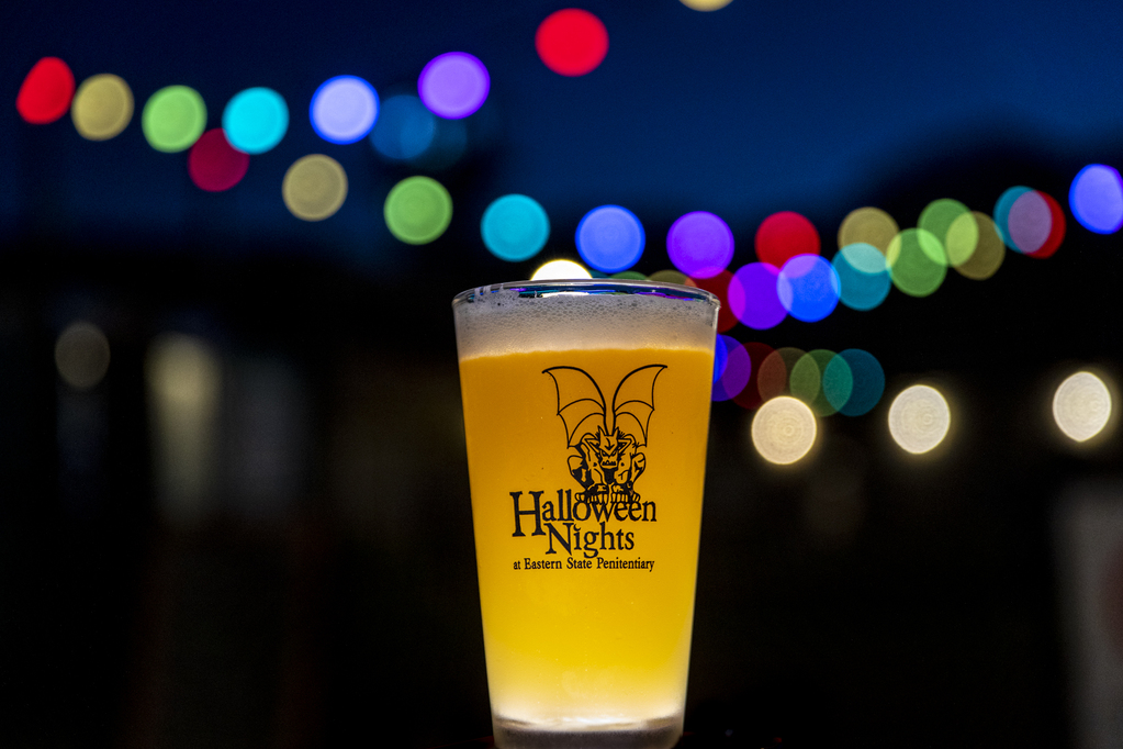 A photo of a glass of beer with the Halloween Nights logo on it. There are colorful lights in the background