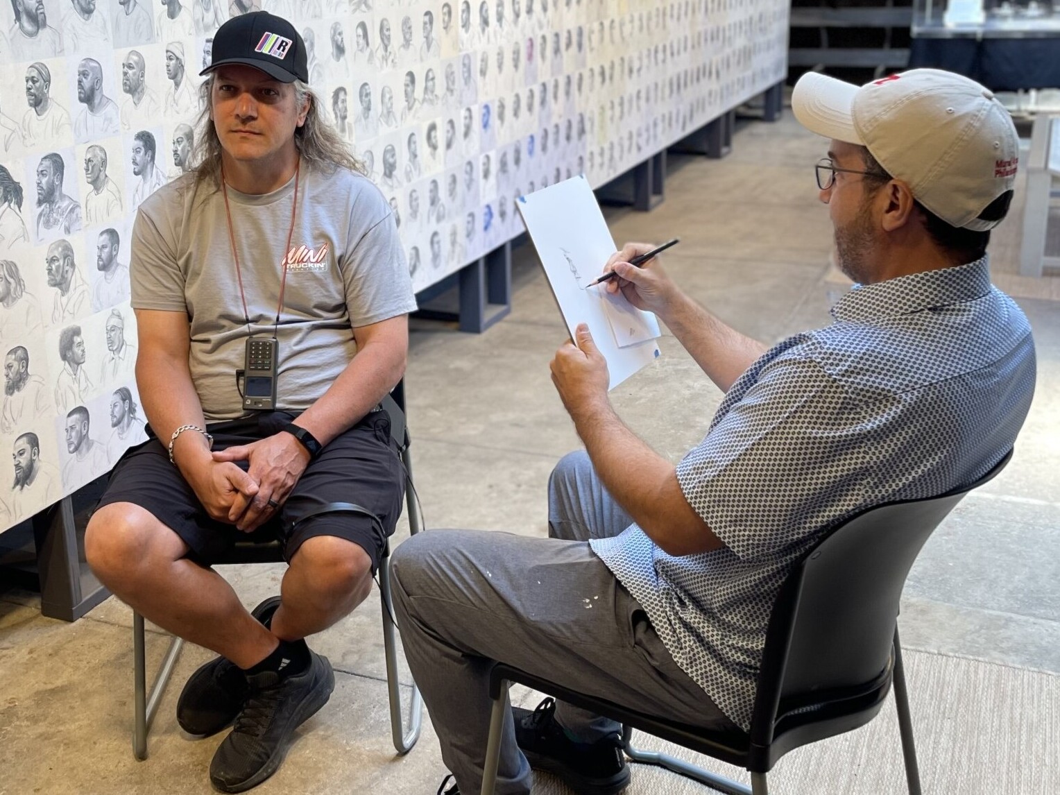 A photograph of artist Mark Loughney sketching a visitor at Eastern State Penitentiary. Behind them is Mark's work Pyrrhic defeat, featuring hundreds of sketches of people.