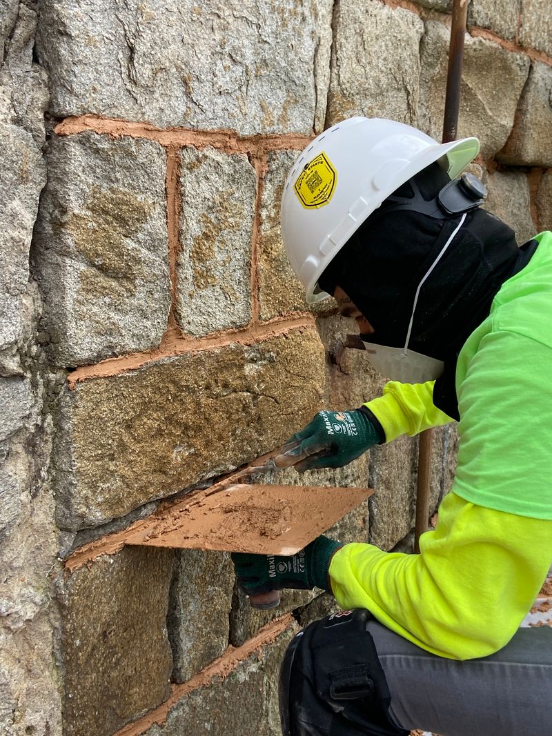 A photo of a LEAD Fellow working at Eastern State Penitentiary. He is wearing a hard hat and completing masonry work on an old stone wall.