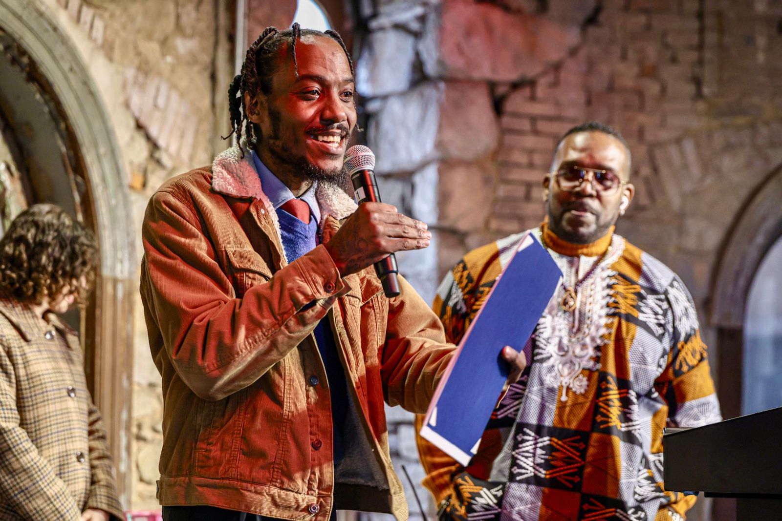A photo of a LEAD graduate talking at Eastern State Penitentiary. The speaker holds a microphone. He is set against aged stone and brick walls.