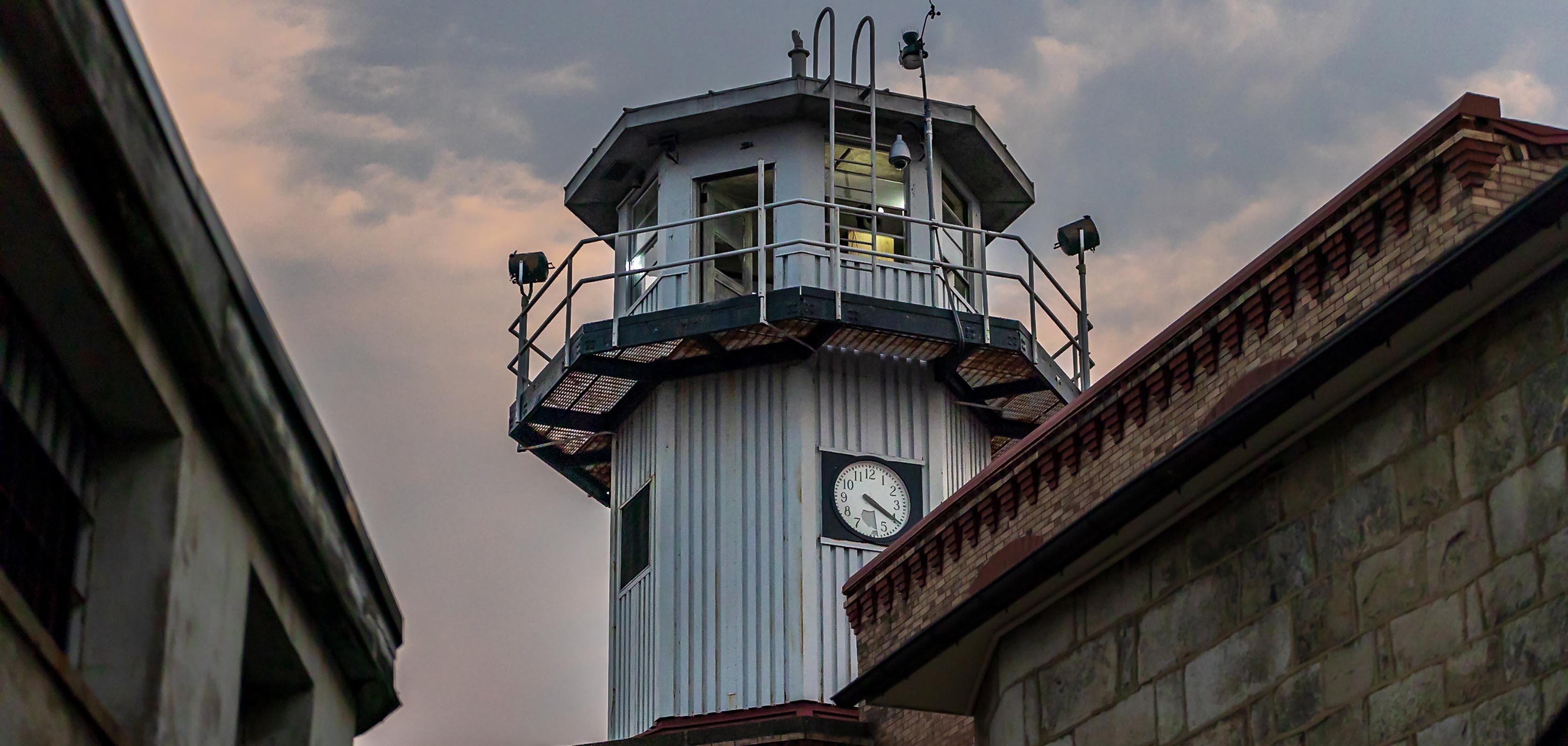 A photo of the central guard tower at Eastern State Penitentiary. The octagonal tower has a clock, metal railing, and lights atop. It’s framed by cloudy skies and flanked by two buildings with brick and stone exteriors.