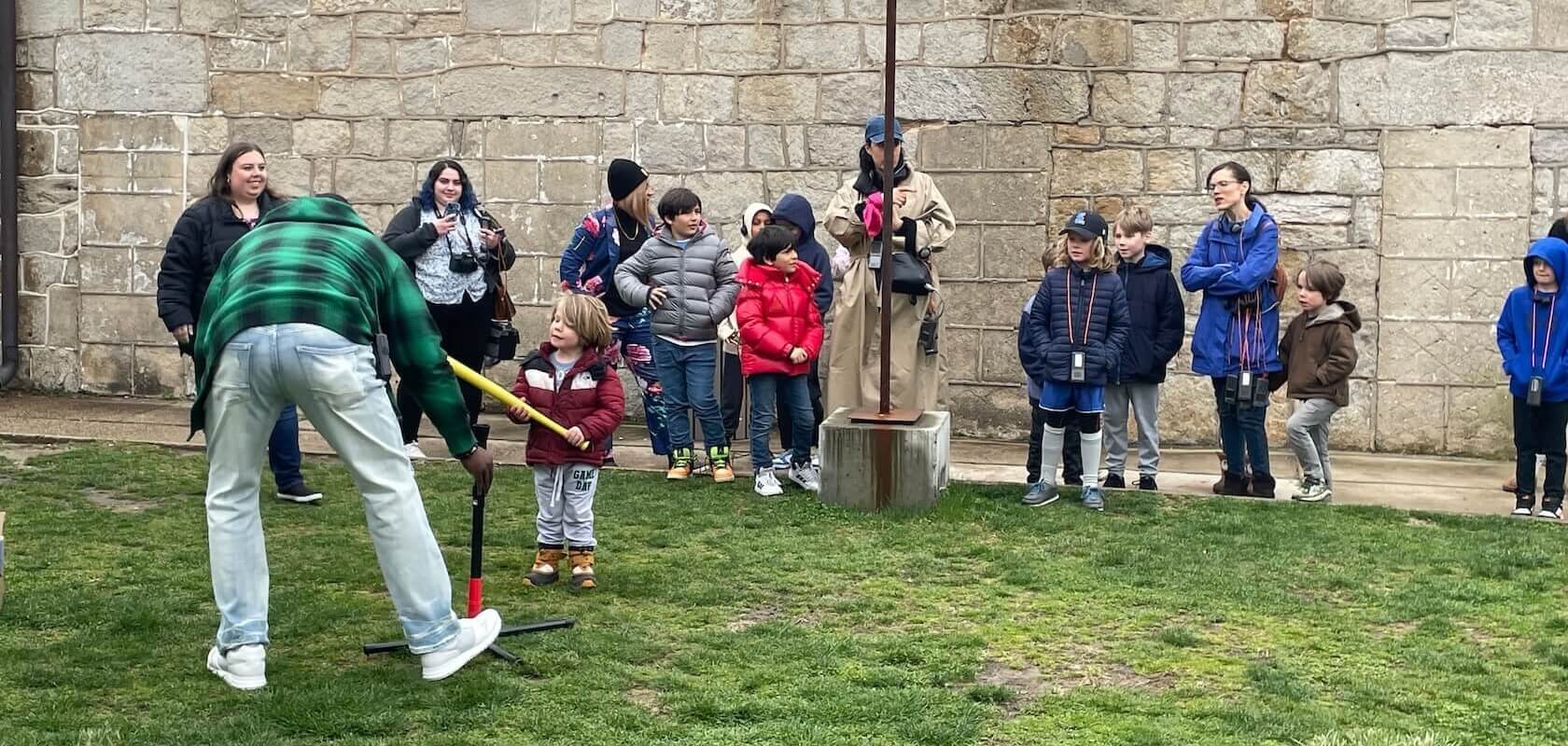 Guests playing baseball at Eastern State Penitentiary. The picture shows a group of adults and children gathered on a grassy area in front of a stone wall. One adult is bending down to help a child with a wiffle ball bat. Everyone is dressed in warm clothing.