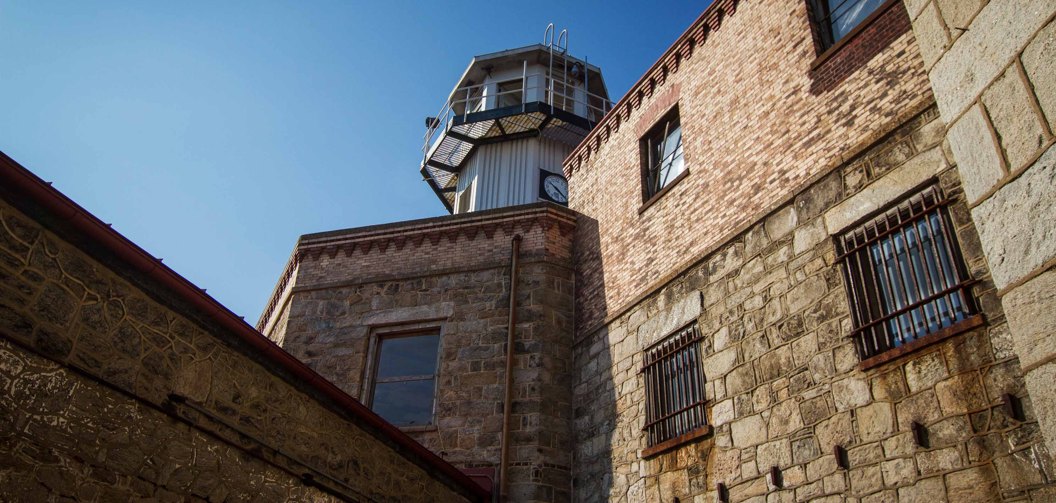 A view of Eastern State Penitentiary's guard tower from below. The exterior of the building is shown, featuring stone and brick walls with barred windows. In the background, there is a prominent watchtower equipped with security features such as metal railings and surveillance equipment.