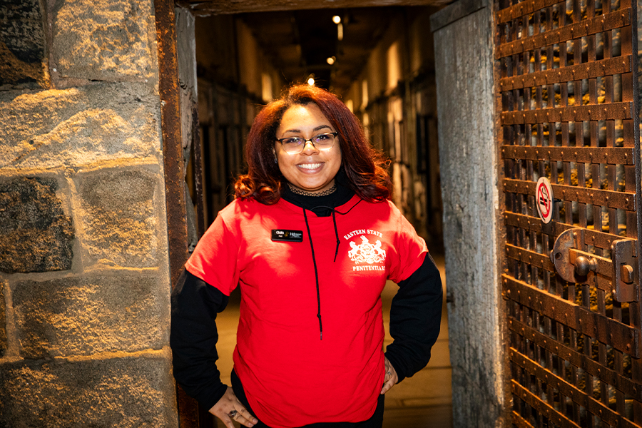 Person in a red staff shirt standing in a stone doorway beside an open barred prison gate inside a historic cellblock.