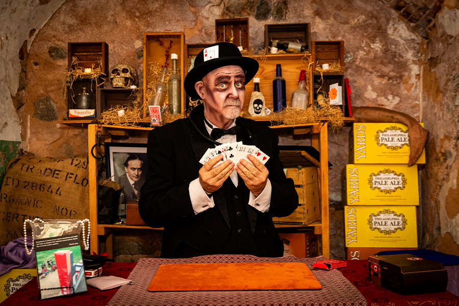 Costumed magician holding a fan of playing cards behind a table in a rustic room filled with vintage props and shelves.