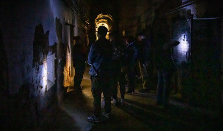 This image captures a dimly lit corridor with several people walking through it, some using flashlights to illuminate their surroundings. The walls are worn and peeling, suggesting the space is aged, abandoned, or historically significant.
The corridor extends into a brighter area in the distance, possibly an exit or another room, creating a strong contrast between light and shadow. The atmosphere evokes a sense of exploration, mystery, or historical preservation, possibly indicating a guided tour or an investigative walk through an old prison, asylum, or similar site.
